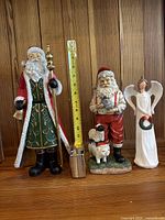 Three festive resin figurines (two Santa and one angel) placed side by side against wood panel background, showing size comparison with measuring tape from 9 to 15 inches.