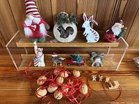 Christmas ornaments displayed on a wooden tabletop with clear shelves, showing a gnome, squirrel plaque, white rabbit, Santa, Lady Liberty holder, dog, red car with tree, silver bells, walnuts, and wooden reindeer sleds.