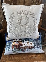 Front view of stack of two decorative holiday pillows on chair, embroidered beige pillow on top, winter village print pillow below