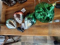 Four assorted metallic holiday ornaments lying on wooden surface, including a large green cut-glass style ornament and three tear-drop shapes in silver and copper.