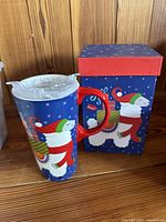 Double-walled insulated glass travel mug with holiday scene design featuring snowman in red scarf, green mittens, blue background with snowflakes, next to matching decorative box.