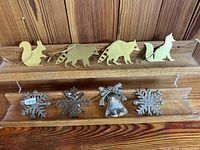 Front view showing 4 gold-tone animal-shaped metal ornaments; squirrel, two raccoons with cut-out stripes, and a cat on a wood holder; below shelf holds 3 silver-tone ornamental pieces: two snowflakes and a bell with bow.