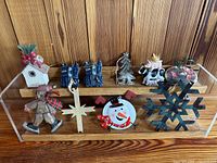 Front view of 10 assorted Christmas ornaments arranged on a wooden display shelf, showing variety of colors and designs including birdhouse, boots, snowflake, and snowman.