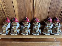 Set of 6 identical resin fireman ornaments lined up on a wooden surface, each holding a coiled fire hose and wearing red helmet with number 25.