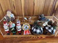 Top view of all 11 Christmas ornaments arranged on a wooden surface with a clear tray, showing the full set including nutcracker ornaments, buffalo plaid jingle bells, gold stars, figurines, and ribbon ornament.