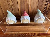 Front view of three ceramic gnome figurines placed on a transparent stand against wooden panel background, showing hats, noses, and beards.