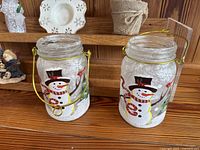 Two glass candle holders painted with snowmen wearing black top hats and scarves, each with a gold metal handle, placed on a wooden surface.
