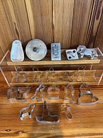 Photo showing all 19 metal cookie cutters arranged on a wooden surface with a clear holder, displaying various whimsical and holiday-themed shapes such as gingerbread men, animals, hats, and cups.