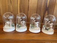 Set of 4 clear plastic domes on white bases with winter scenes including deer and pine cones inside, all displayed on wooden surface.