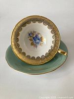 Top view of teacup showing gold filigree lace design and central blue rose bouquet inside the cup, sitting on matching turquoise saucer with gold rim.