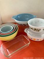 Photo showing colorful nested Pyrex bowls with two patterned bowls (blue with grapevine and white with green floral) and clear glass baking dish on orange surface.