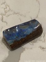 Top view of polished boulder opal specimen showing the vibrant blue opal vein against the brown matrix on a light marble surface.