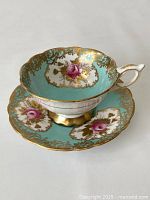 Side view of turquoise teacup on matching saucer showing gilt floral design and pink roses inside cup and on saucer, scalloped edges, and white handle with gold trim.