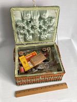 Open wicker sewing basket showing satin-lined lid with needles and pins secured, visible contents include spools of thread, scissors, vintage packaging, and assortment of small sewing items inside compartments