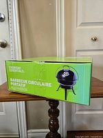Photo showing the Everyday Essentials brand name and the box with the image of a black, circular, portable charcoal barbecue grill with handle on lid and three legs.