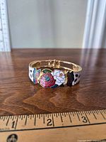 Front view of vintage gold-tone bracelet with colorful butterfly and floral enamel designs on wooden surface with measurement scale.