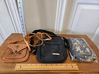 Three small bags displayed on a wood surface with measuring rulers showing size; tan leather vintage bag with carved flower designs, black canvas bag, and camouflage bag in wrapping.