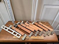 Eight adult sized pant hangers arranged on a wooden table. Seven wooden hangers with metal clips and one white plastic hanger with black clips are positioned together showing hooks and clip details.