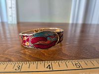 Side view of the gold-tone bracelet with colorful enamel heron design and surrounding floral patterns, placed on a wooden surface with a ruler showing length.