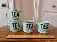 Four light green enamel mugs stacked and arranged on wood surface, showing outside text 'TEA TASTES BETTER OUTDOORS!' in black with metallic rim top edges.
