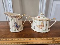 Porcelain creamer and lidded sugar bowl set on wooden surface with measuring rulers behind showing size. Cream color base with butterfly, insect and flower decoration and gold trim on rims and handles.