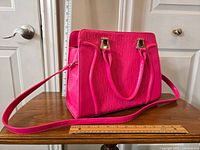 Hot pink handbag resting on wooden table, showing embossed pattern and gold-tone hardware, ruler placed next to it for scale.