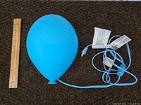Blue balloon-shaped lamp with attached power cord and plug placed on a carpeted surface next to a wooden ruler for size reference.
