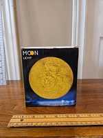 Box of moon lamp showing yellow moon image on front with moon lamp branding. Rulers placed around for size reference.