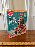 Boxed Klutz Mini Pom-Pom Pets craft kit upright on wood table with ruler next to it showing size, sealed packaging with colorful pom-pom animal illustrations on front.