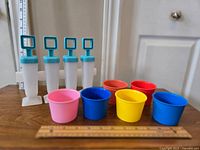 Four turquoise and white popsicle makers next to six colorful plastic snack cups arranged on a wooden surface.