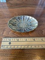 Oval brass trinket dish or ashtray placed next to a ruler indicating approximately 4.5 inches length; shows detailed ornate engraving and aged patina.
