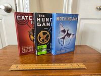 Three hardcover books from The Hunger Games series by Suzanne Collins standing vertically with a ruler in front for scale.
