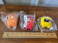 Three colorful monster-shaped dog toys in clear plastic, placed beside a ruler showing their size around 3 inches each.