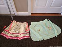 Two vintage aprons laid flat side-by-side: a crocheted cream apron with pink ribbons, and a green sewn apron with floral embroidery.