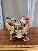 Front side view of four seashell frogs sitting around a seashell table, each holding miniature cards and wearing distinct colored caps.