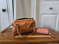 Photo showing vintage tan leather handbag with handles, front emblem, and Maurice's pink wallet on wooden surface with measurement rulers for size reference.