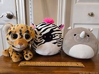 Three plush toys on wooden surface: Beanie Boo Cheetah, Squishmallow Zebra, and Squishmallow Rhino with ruler for size reference