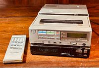 Overview of Panasonic PV-9000 portable VCR on top paired with Panasonic PV-A860 VHS player and original Panasonic remote control on wooden surface.