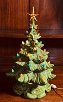 Classic view of the ceramic Christmas tree showing the green color, textured branches, multicolored bulbs, and amber star topper. Plug cord shown for power.