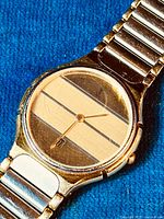 Close-up photo of the round watch face with striped gold-tone dial showing linear patterns and gold-tone hands. Date display window at 6 o'clock is visible. Shows some wear on the watch crystal.