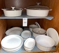 Cabinet shelf holding assortment of white Corning Ware ceramic bakeware dishes and glass lids, showing overall lot contents