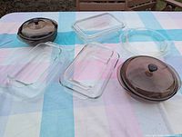 Photo showing all glass cookware laid out on tablecloth, including clear rectangular Pyrex casseroles, round tinted brown Pyrex casseroles with lids, clear pie plate, and round tinted brown cake pan.