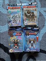 Photo showing front side of four unopened Gundam action figures in blister card packaging placed on blue carpet.