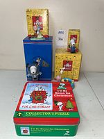 Photo showing four Snoopy collectible figures atop and beside their original yellow themed Peanuts boxes and the puzzle tin with Christmas artwork on the front.