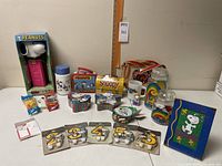 Full collection of Snoopy and Peanuts items arranged on table including Pez dispenser, lunch tins, storage jars, mug, picture frame, birthday candles, and shoulder bag.