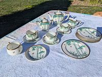 Full tea set displayed on white cloth outdoors showing teapot, creamer, sugar bowl, 6 plates, and 4 cups with saucers, all with green bamboo design and gold trim.