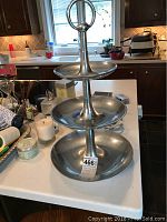 Full view of a heavy metal 3-tiered server standing on a kitchen counter. Shows the entire tiered server with ring handle on top and three progressively larger circular trays from top to bottom.