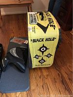 Black Hole four-sided foam archery target block with caution label and black Easton quiver leaning against it on hardwood floor.