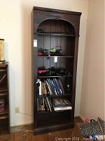 Front view of tall dark wood bookcase with books and items on shelves, showing ornate top trim and six shelves.