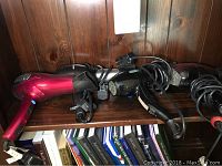 Photo showing four blow dryers with attached cords placed on a wooden shelf.